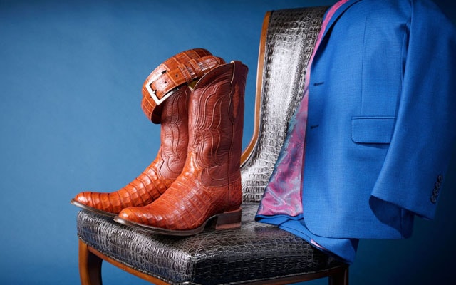 A pair of the Tremaine Cognac 12” Men’s boots, the Genuine Nile Crocodile Belt in brandy, and a sports coat sitting on a gray caiman chair.